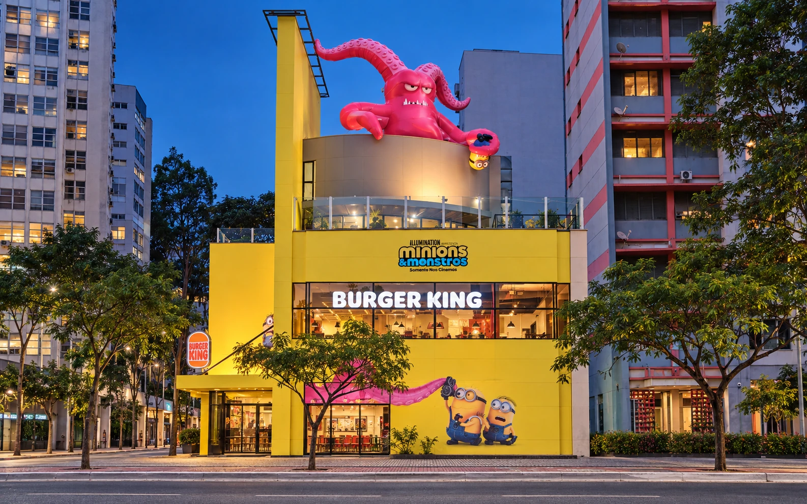 Fachada do Burger King na Avenida Paulista envelopada para Minions e Monstros, com monstro inflável gigante no topo ao anoitecer