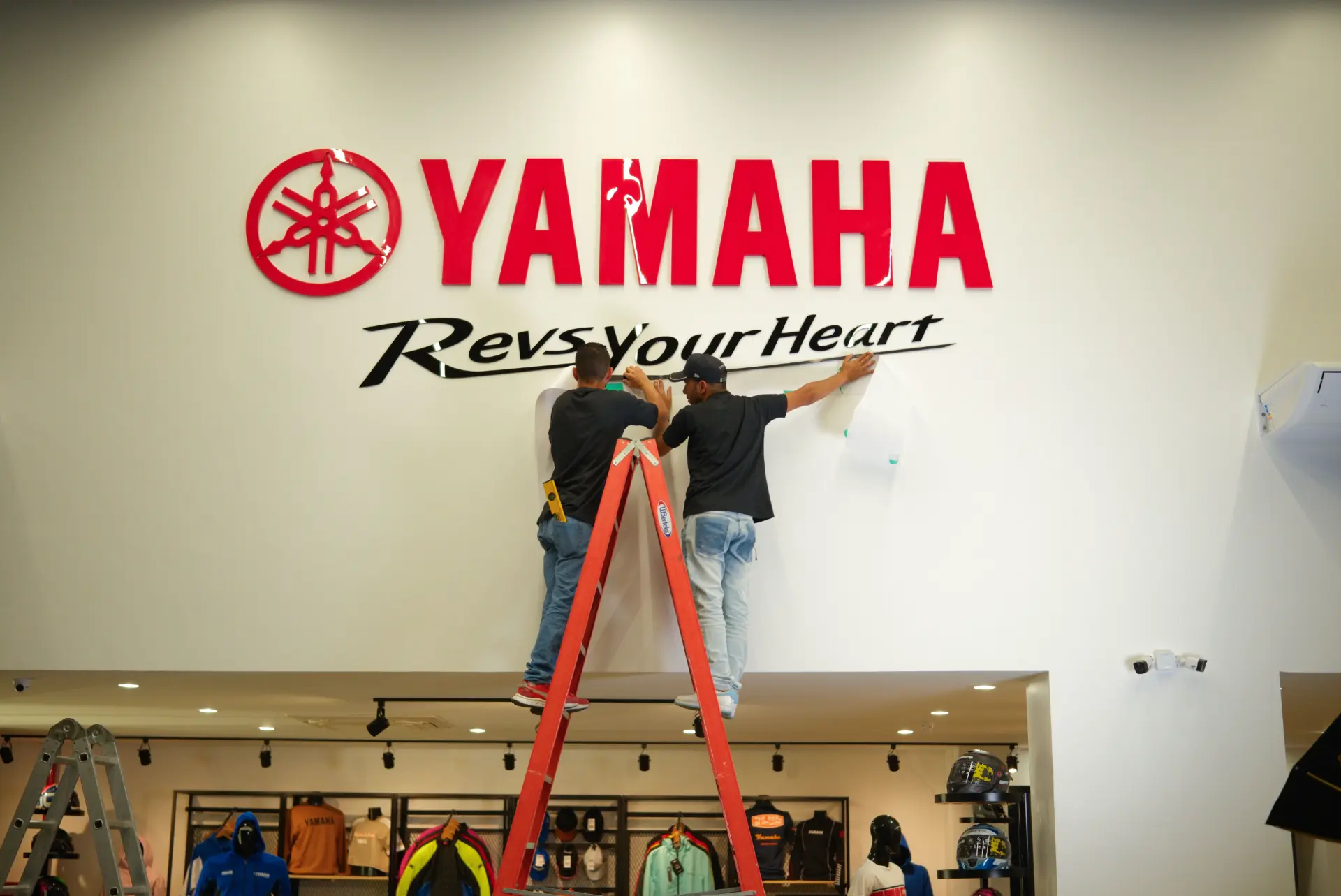 Instaladores aplicando o letreiro Revs Your Heart sob o logo Yamaha vermelho na parede da loja